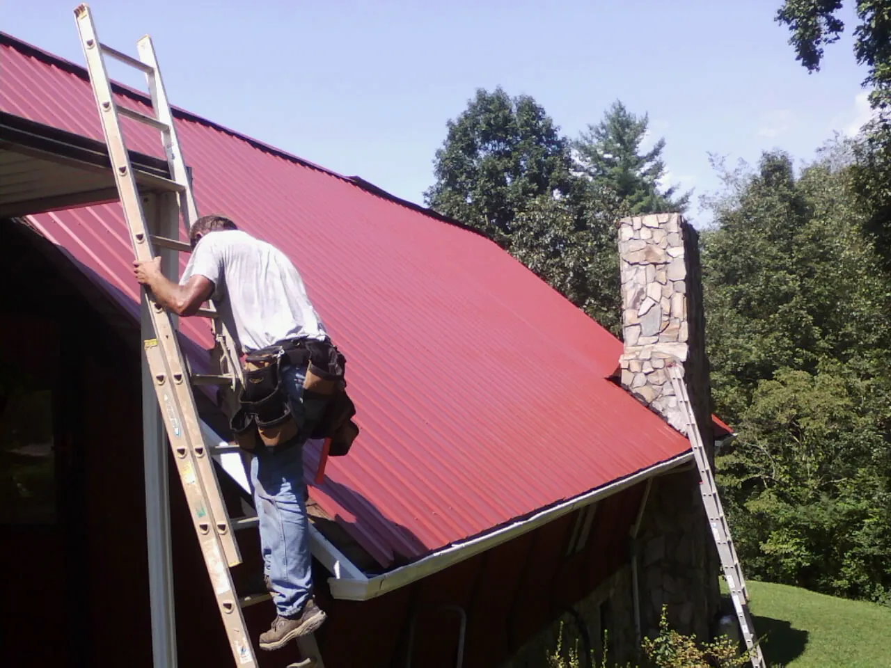 Roofer on a ladder beside a red metal roof and stone chimney — Soffit & Fascia Repair by Roofing Brothers in Brookline